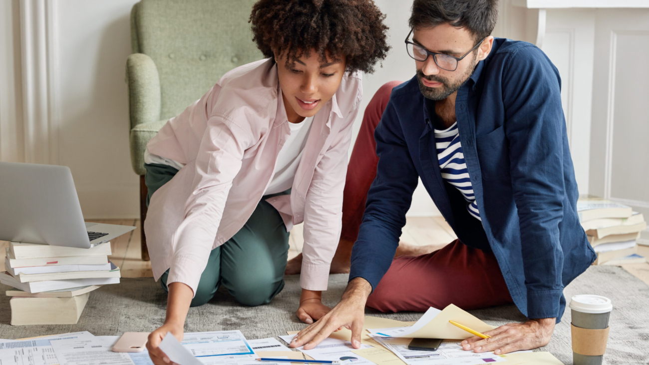 Horizontal shot of multiethnic investors study investment portfolio together, surrounded with paper documents, books, modern technologies and takeaway coffee, have full time job, work on weekend